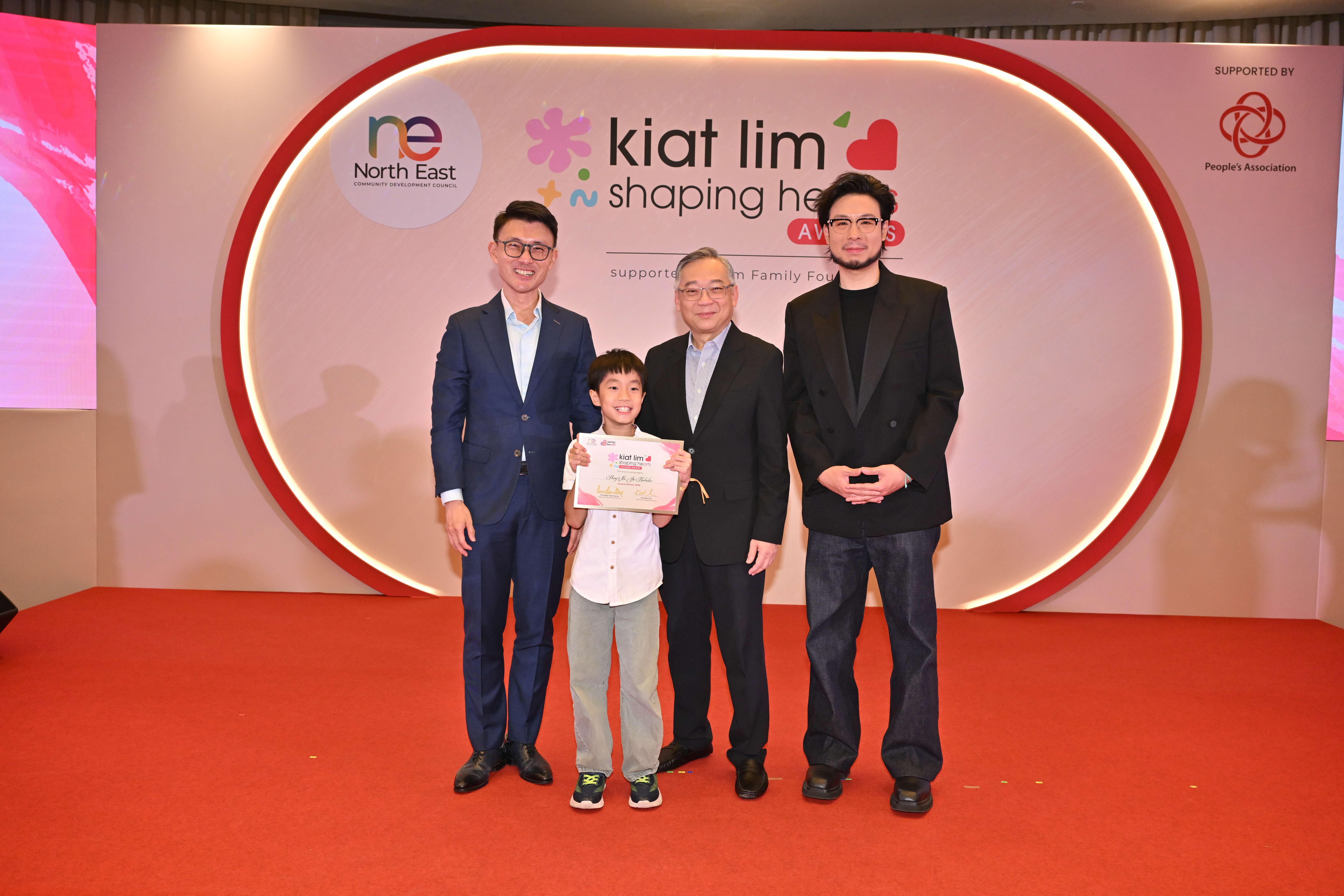 Award recipient standing on stage with the presenter, holding their certificate and posing for a group photo during the award ceremony, with a backdrop reading ‘Kiat Lim-Shaping Hearts Awards'.
